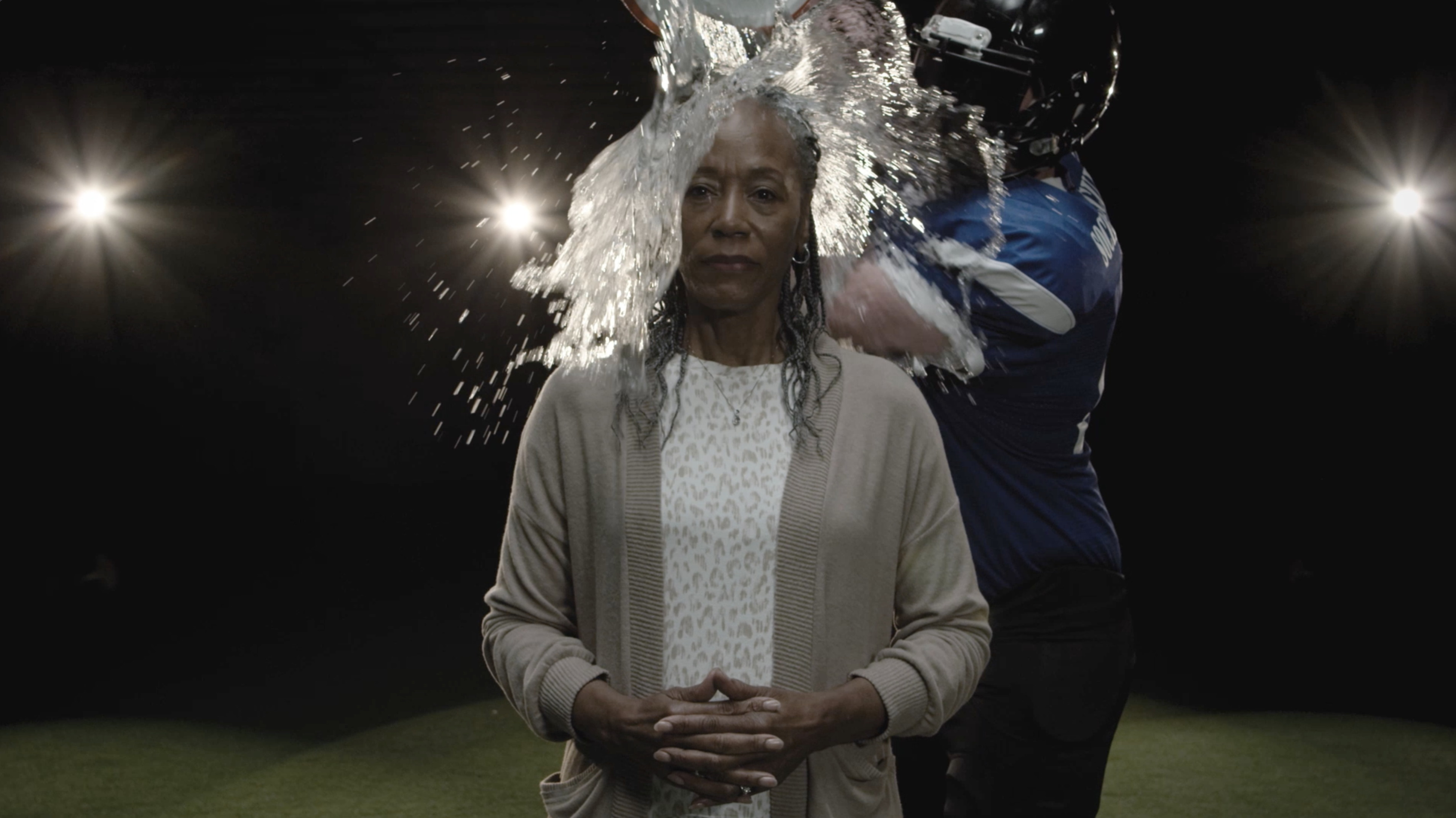 A woman stands looking into the camera with her hands together as a football player dumps a container of water onto her head